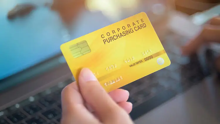 A hand holds a corporate purchasing card over a laptop keyboard